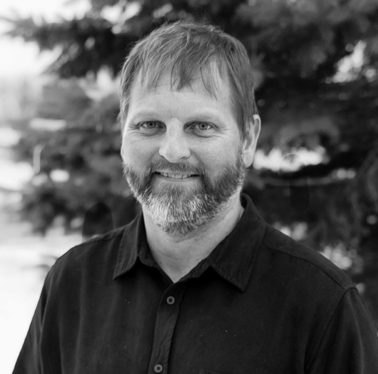 Smiling man with a light beard in a black button-down shirt, outdoors with trees in the background.