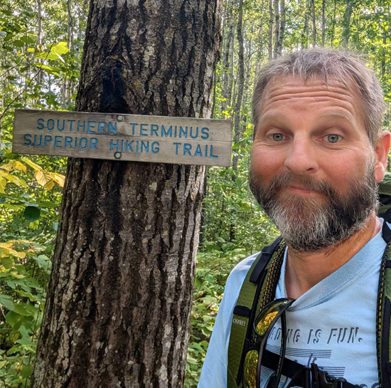 Hiker with beard taking a forest selfie beside a wooden sign reading 'southern terminus superior hiking trail' - ironistic