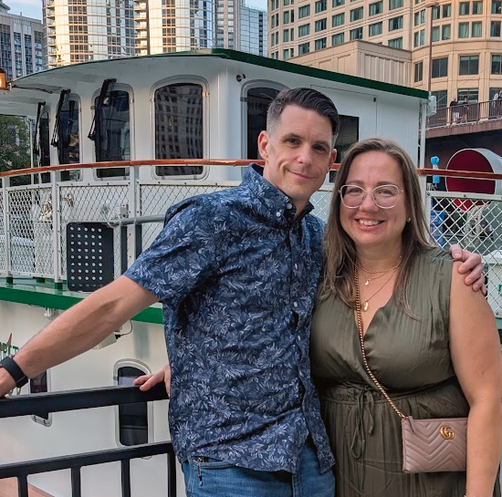 Smiling couple posing arm-in-arm on a marina railing with a white boat and city buildings behind them; man in a blue floral shirt, woman in a green dress with a pink purse. - ironistic