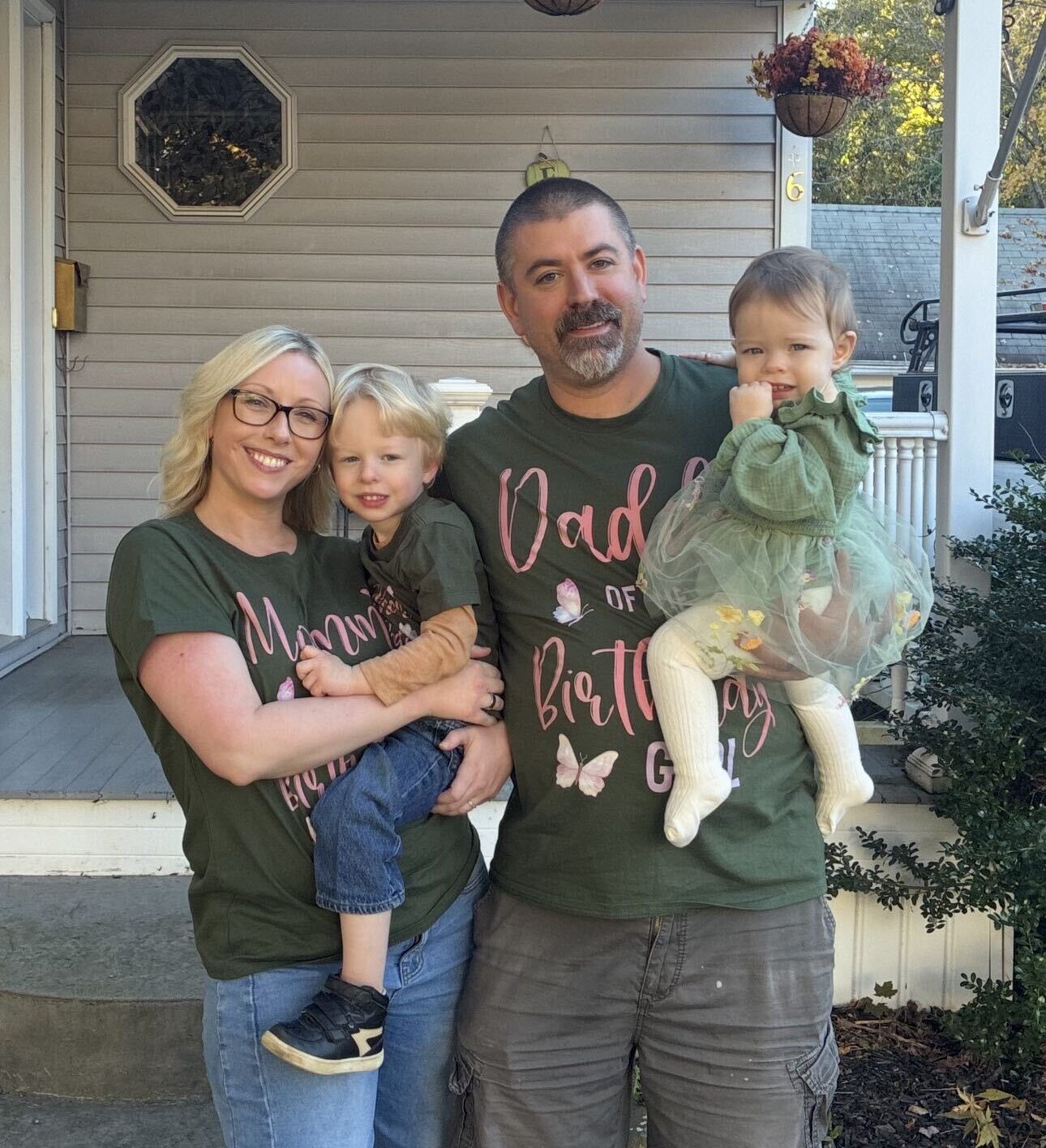 Family of four posing on a front porch: mom holding a toddler, dad holding a baby, all wearing matching green shirts and smiles. - ironistic