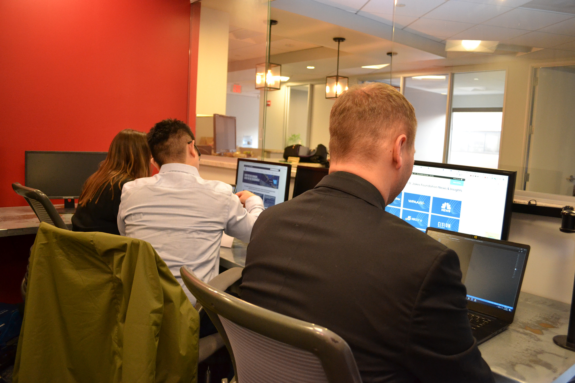 Open-plan office with people at desks using computers; red wall on the left and glass partitions in the background.