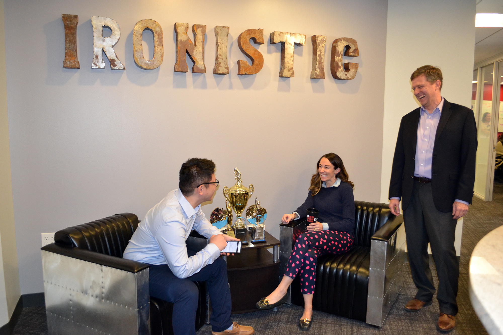 Office-lobby - ironistic Office lounge with three coworkers around a coffee table displaying trophies; one man stands and smiles in a suit, while two sit on leather sofas. - ironistic