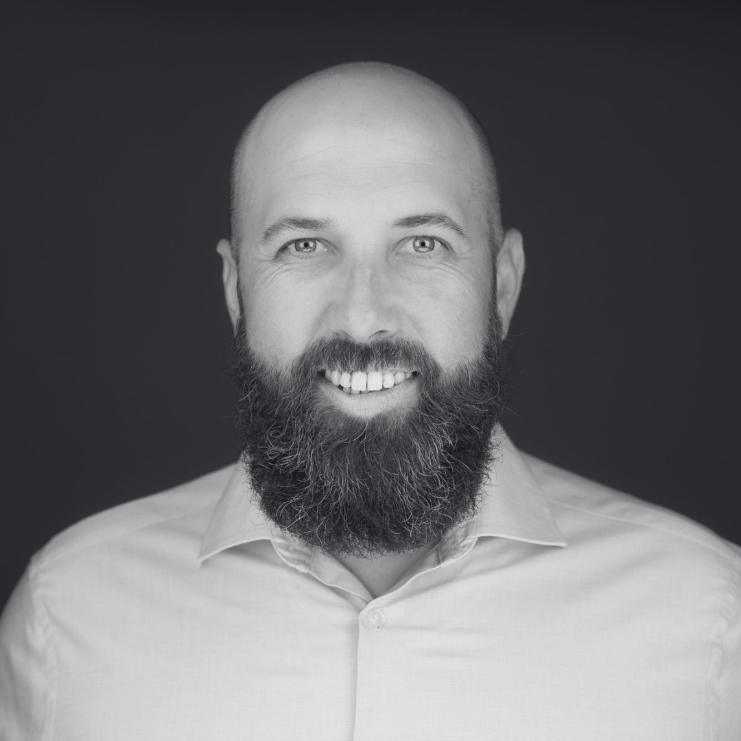 Head-and-shoulders portrait of a smiling man with a full beard and a shaved head, wearing a light button-up shirt against a dark background.