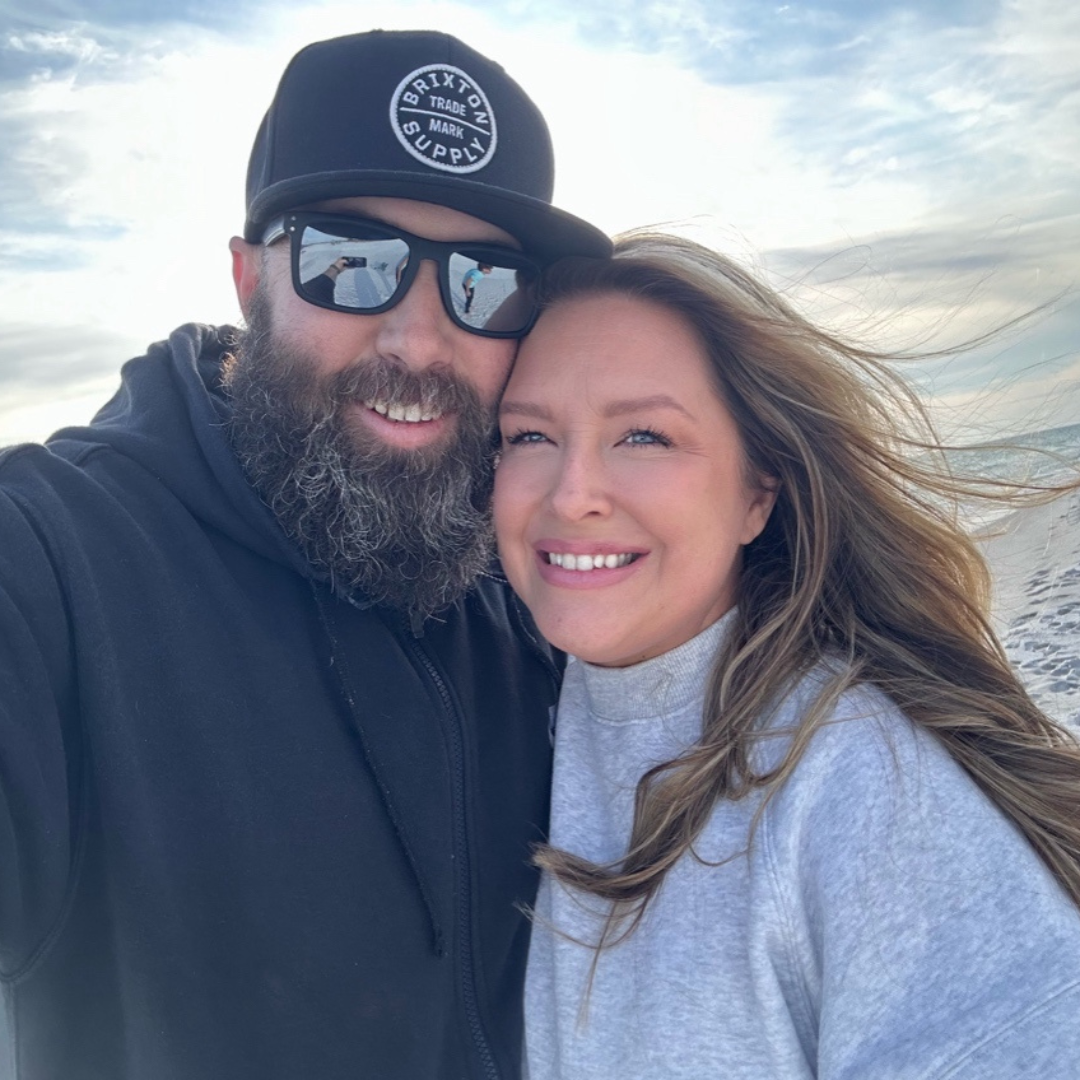 Smiling couple taking a selfie on a windy beach, man with a beard in a black hoodie and cap, woman in a gray sweater beside him.